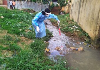 Ein Person in Schutzkleidung beim Entnehmen einer Wasserprobe mittels eines Geräts aus einem Wasserrinnsal, in welchem auch diverser Haushaltsmüll herumliegt.