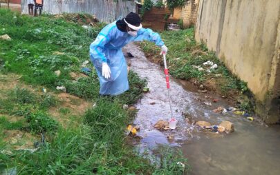 A person in protective clothing taking a water sample using a device from a small stream in which various household waste is lying around.