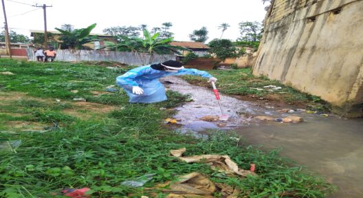 Ein Person in Schutzkleidung beim Entnehmen einer Wasserprobe mittels eines Geräts aus einem Wasserrinnsal, in welchem auch diverser Haushaltsmüll herumliegt.