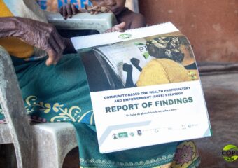 The hands and lap of an older seated woman holding a spiral-bound document. The document includes the text “Report of Findings” and a sentence in Igbo language.