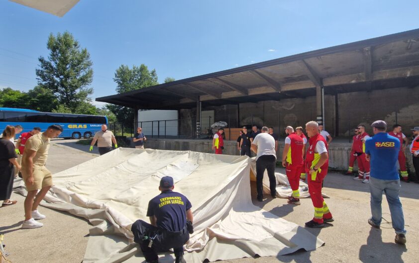 Exterior shot: Several people are setting up a large tent, among them some wearing ASB T‑shirts and others in work wear (red vests and trousers with reflective strips).