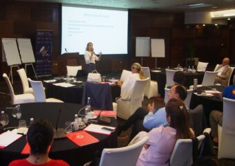 IIn a room, people are sitting at round tables with documents and microphones. A woman stands in front of a PowerPoint presentation and speaks into the microphone to the attendees. Flipchart boards are located next to the speaker's podium.