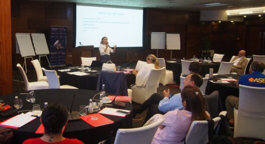IIn a room, people are sitting at round tables with documents and microphones. A woman stands in front of a PowerPoint presentation and speaks into the microphone to the attendees. Flipchart boards are located next to the speaker's podium.